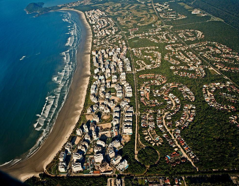 Vista aérea de praia com ondas, arranha-céus à beira-mar e bairro em meio à mata, em tons de azul, areia e verde.