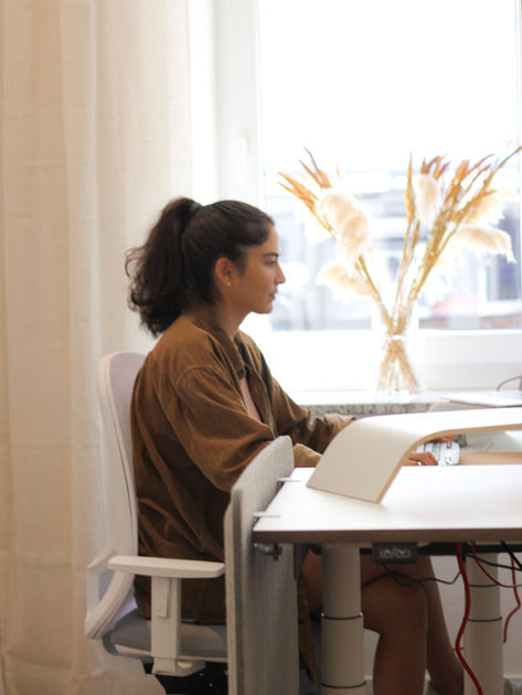 Bild einer Frau mit schwarzen Haaren, die zu einem Zopf gebunden sind, während sie an einem Tisch mit Fenster im Co-Working-Space von Heimatoffice 26 in München sitzt.