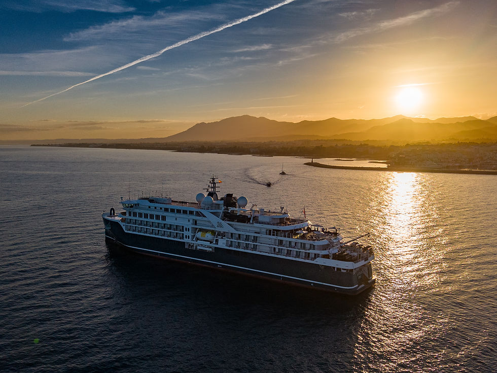 Large cruise ship on calm sea at sunset, golden light on water, mountains and coastline in the distance.