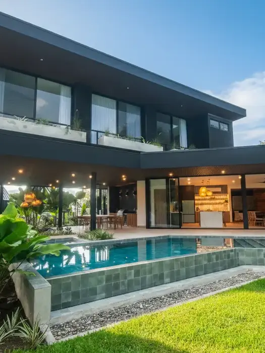 Evening view of Xanadu Arenal Villa featuring a glowing blue infinity pool, a white waterslide, and a modern cantilevered terrace with warm wood ceilings and designer lighting.