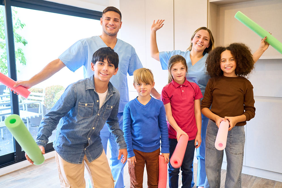 Group of cheerful children participating in pediatric therapy activities with two smiling therapists wearing blue scrubs in a bright rehabilitation setting