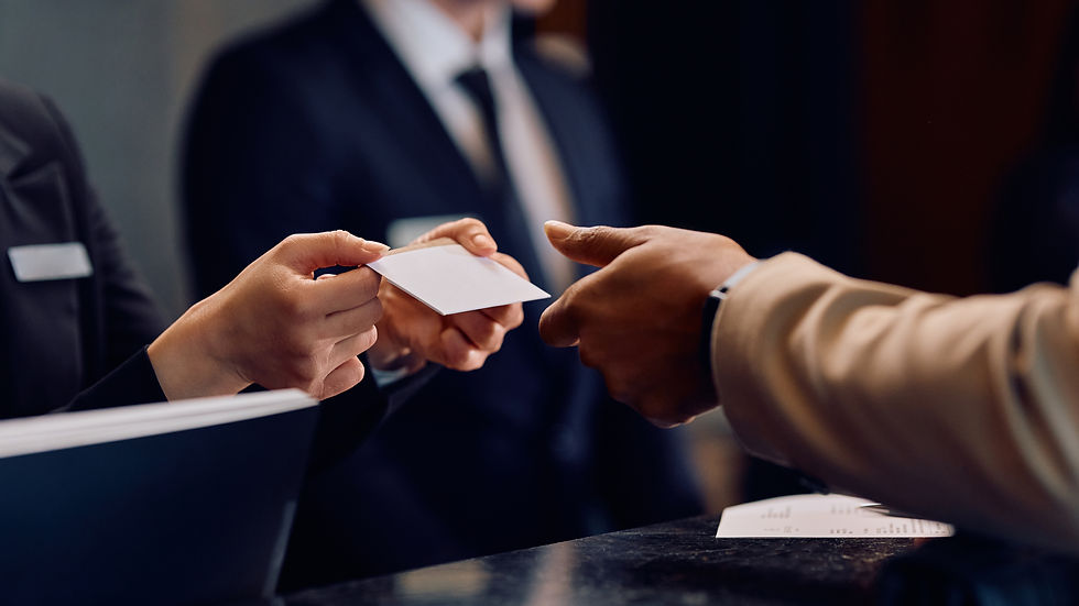 Hands exchanging a card over a hotel reception desk. One person wears a black suit with a name tag. Professional and formal atmosphere.