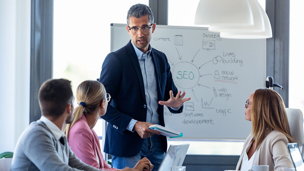 Man in a suit presents at a meeting in front of a whiteboard with "SEO" diagram. Attendees listen attentively. Bright office setting.