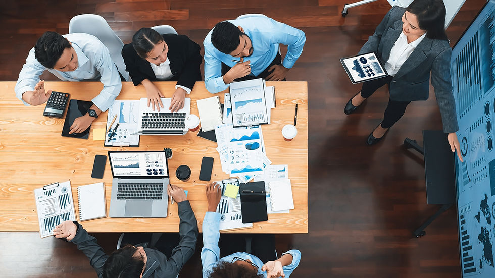 Top view of a team meeting with six people around a wooden table. Laptops, charts, and coffee cups are present. A person stands, presenting data on a screen.