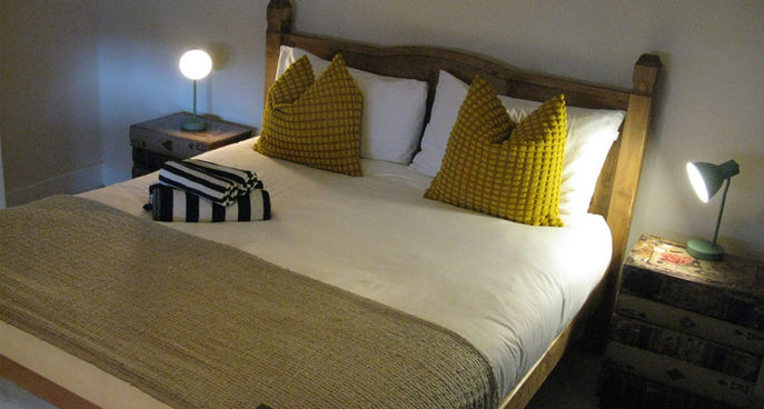 Clean double bedroom with wooden bed frame, yellow cushions, bedside lamps and folded towels.