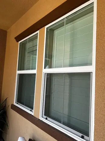 Two double-pane windows with white frames are set in a tan stucco wall, featuring brown trim. A plant is partially visible in the lower left corner—an ideal spot for residential pressure washing in Riverside or Menifee.