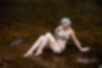 Woman reclining in a shallow river, wearing a colorful bikini and smiling softly while looking down, exuding serenity and confidence like a river goddess.