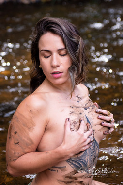 Woman standing in the river with eyes closed, covering her chest with her hands, her tattooed arm visible as she radiates calm confidence and natural beauty.