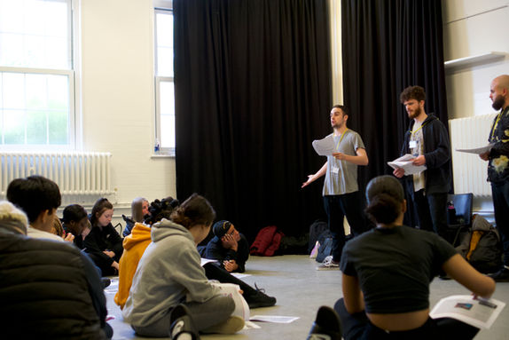 Inside a large drama studio, 3 Voicebox facilitators leading a workshop to a group of young people. The young people are sat on the floor listening.
