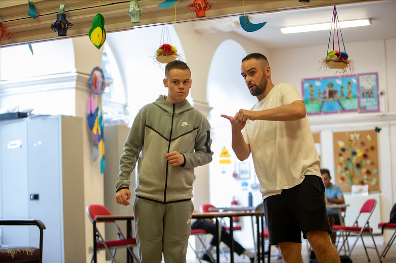 A photo of Voicebox's Project and Outreach Director Jack Tricker, talking to a young participant. They are standing inside a large hall with tables and chairs set out. They both look in the same direction, with Jack gesticulating and the participant concentrating on what is being said.
