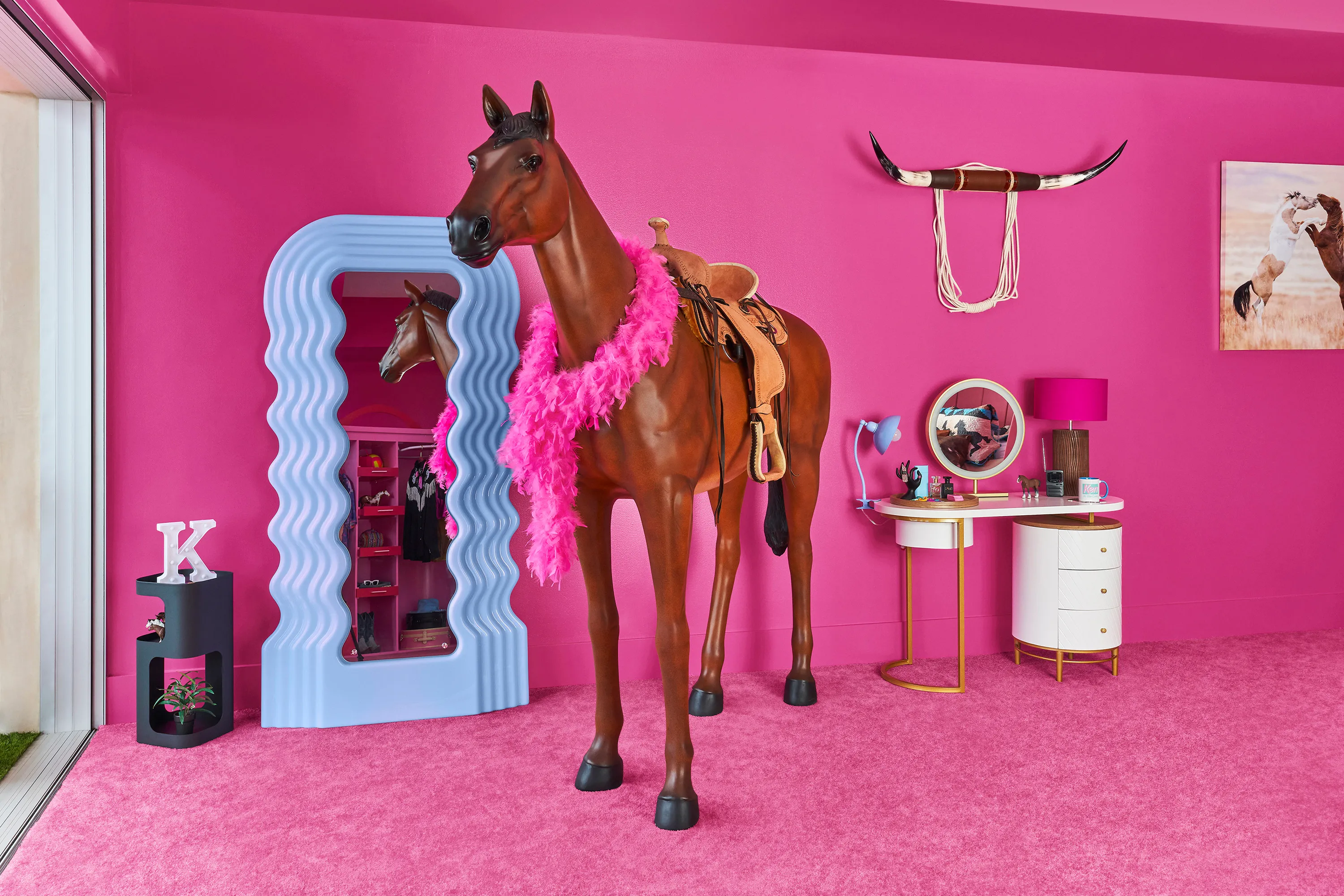 A plastic toy horse in a room of a dolls house. The room's walls and floor are hot pink. On the horse is a feather boa in the same hot pink. Behind the horse is a doll-size mirror, a dressing table, and a table with a letter 'K' propped up on it.