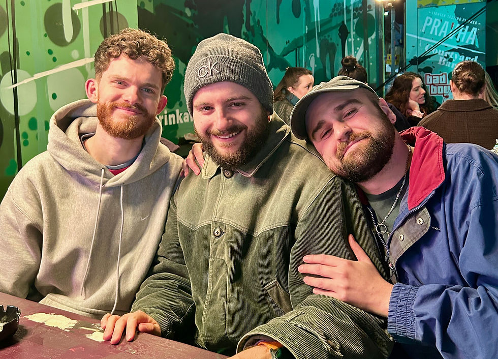 A photo of Voicebox directors Andy, Fredi and Jack sat a table in a busy outdoors bar, smiling at the camera. Andy is on the left, Fredi's in the middle, and Jack is on the right. Jack is leaning into Fredi and has his arms around him.