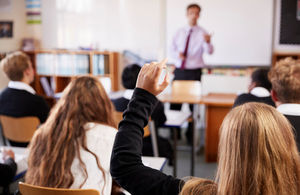 A photo from the back of a school classroom. A student's hand is raised. In the background is a teacher standing, out of focus.