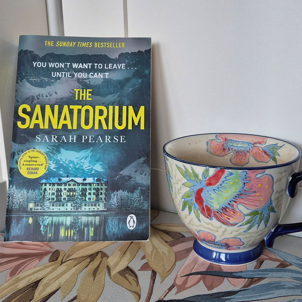 Book "The Sanatorium" by Sarah Pearse with a snowy mountain cover, placed next to a colorful floral mug on a patterned surface.
