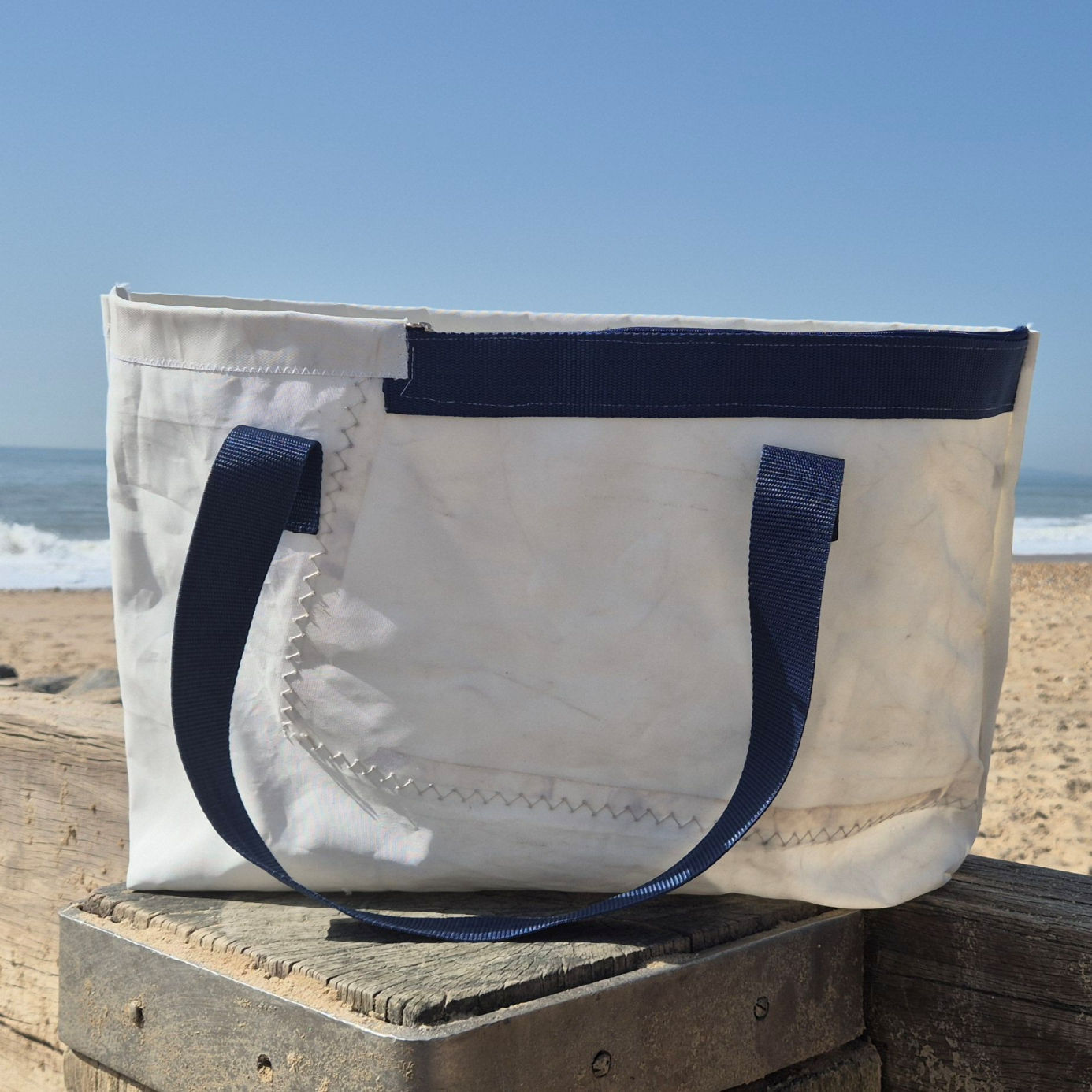 White sail bag with blue handles and blue stripe at top front on the beach