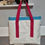 Thumbnail: White Sail Bag on grey background. Hanging from hook via Raspberry coloured handles, light blue finish on top.