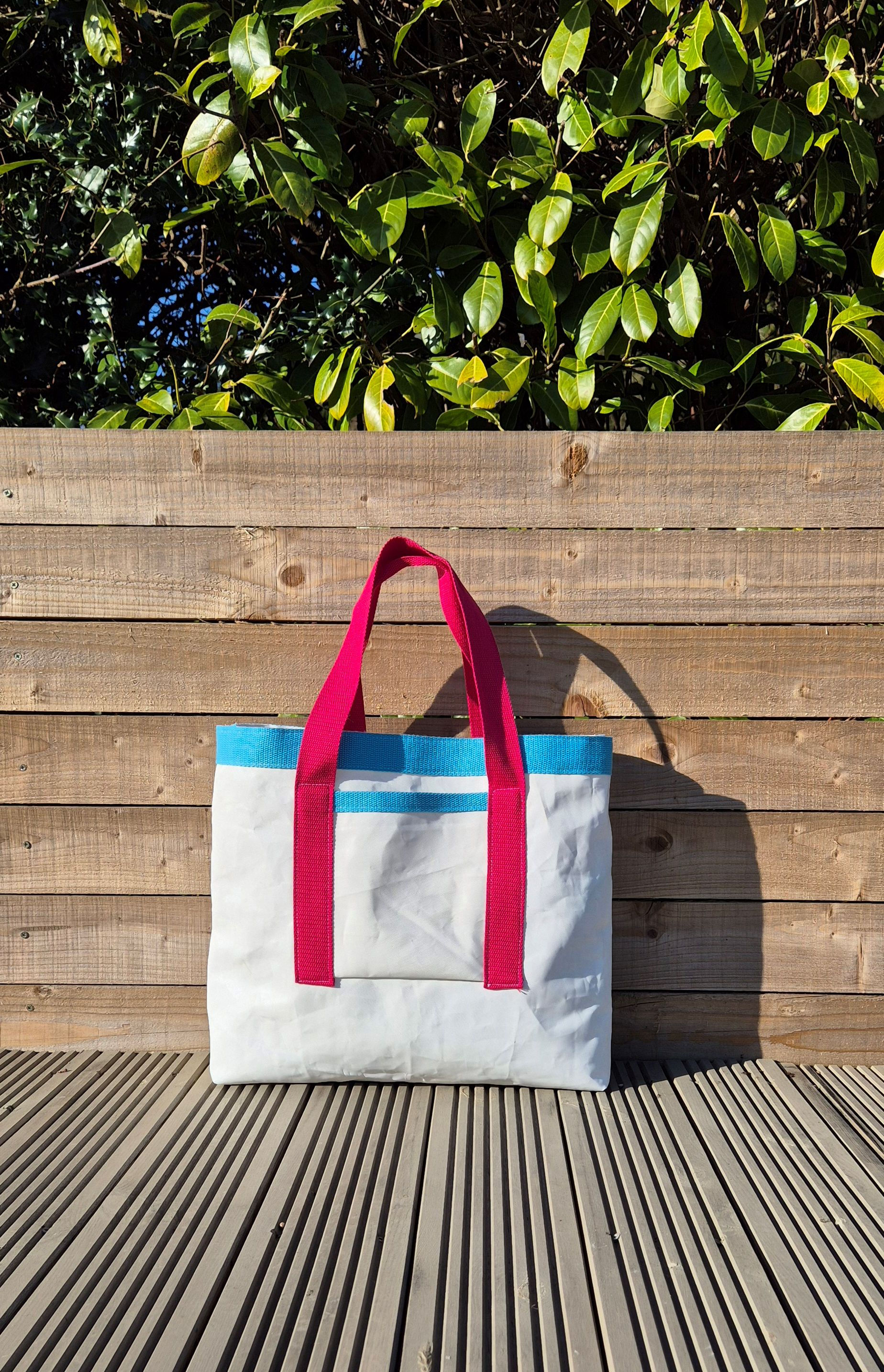White Sail Bag with Raspberry coloured handles, light blue finish on top and front pocket. fence background.