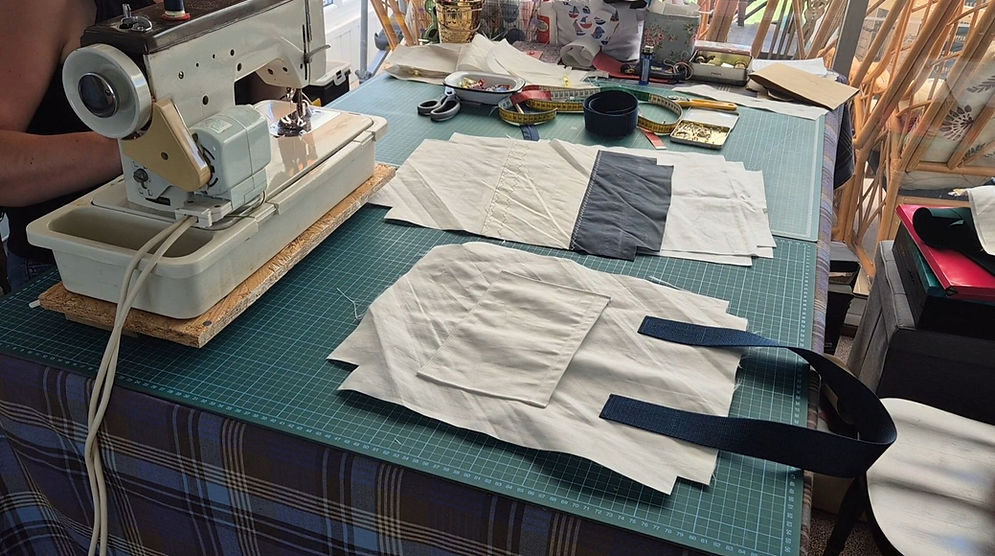 busy table with sewing machine, green cutting covers, some sailcloth pieces, scissors, measuring tape. Person sitting to the left behind maching