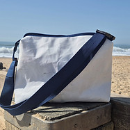 White rectangle Sail Bag with blue zip on top and blue Handle lying across, sitting on a pole at the beach.