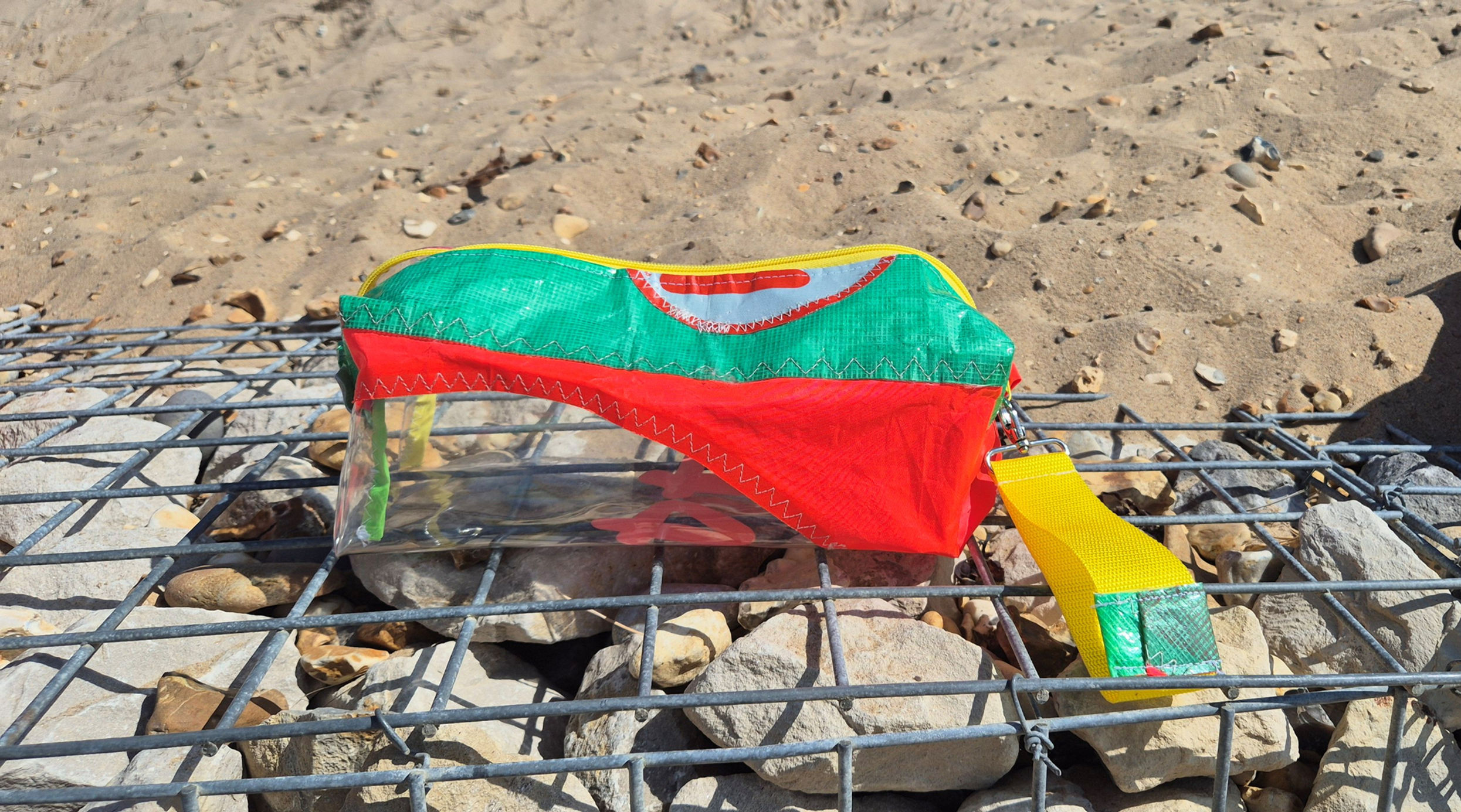 A clear pencil case with a red, green, and yellow fabric top. The case has a yellow zip and a yellow fabric handle.