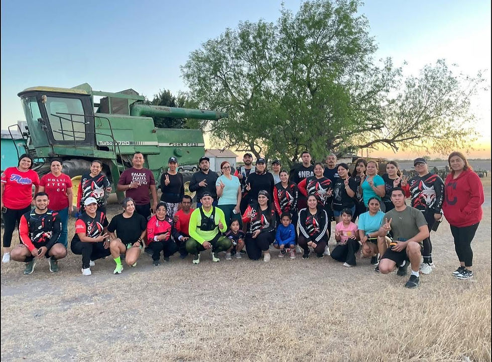 Group of people posing outdoors near a green harvester and tree at sunset, wearing colorful sportswear, expressing joy and camaraderie.