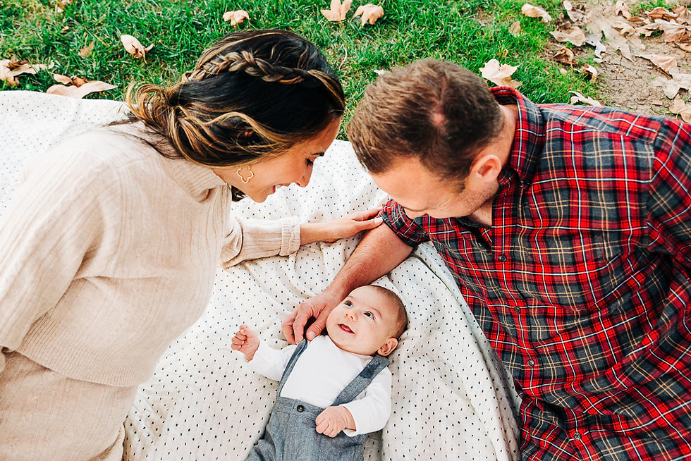 Adults and a baby lie on a picnic blanket on grass, smiling at each other. The woman wears beige, the man red plaid. Leaves scattered around.