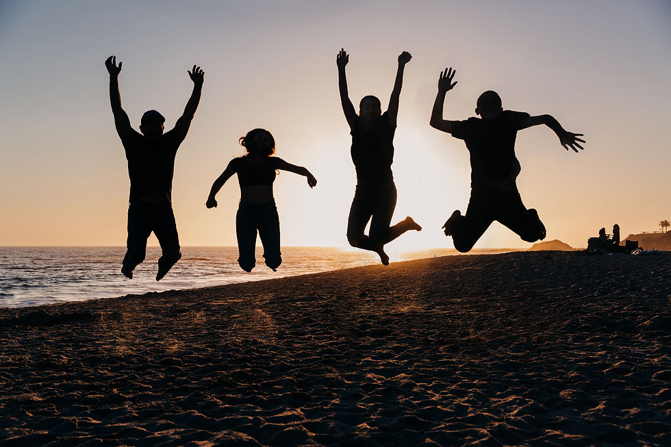 Silhouetted group of four people jumping on a beach at sunset, exuding joy. Ocean and distant figures in background.