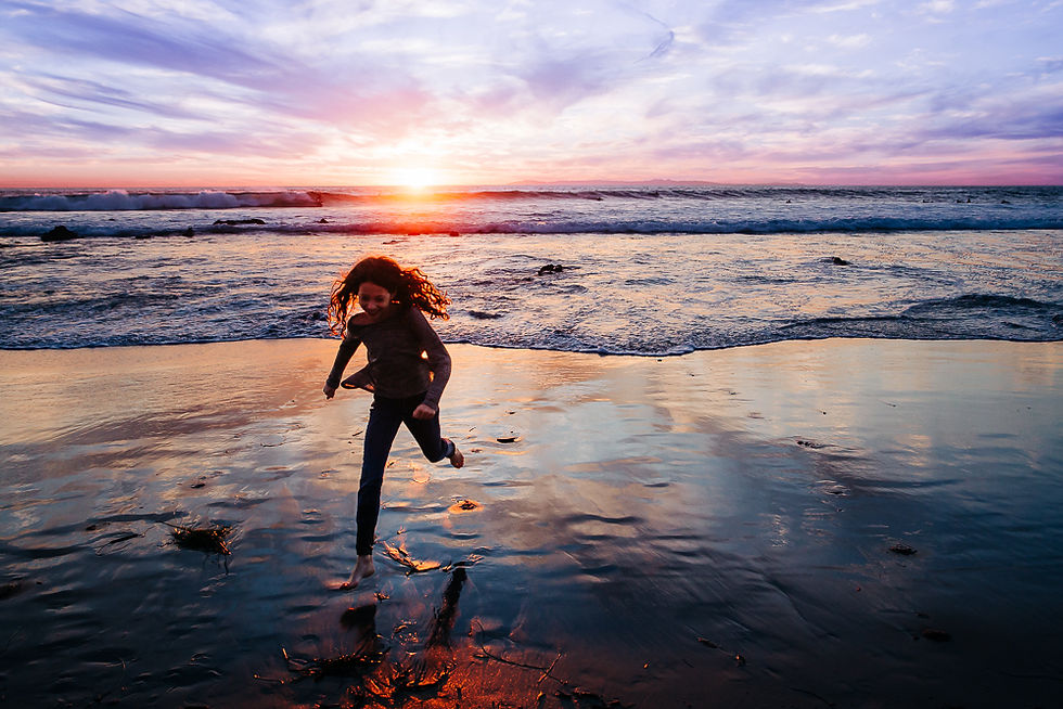 Child running joyfully on a wet beach at sunset, with vibrant purple and orange skies reflecting on the water. Waves gently crash in the background.