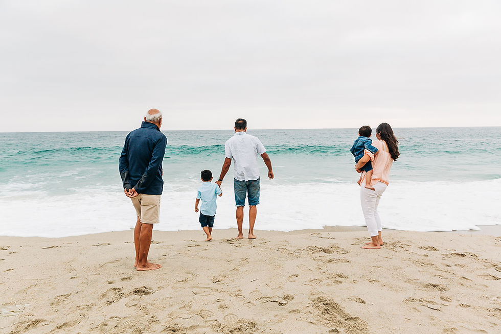 A family of five stands on a sandy beach, facing the ocean. Two adults hold children; one adult stands nearby. The mood is calm and serene.