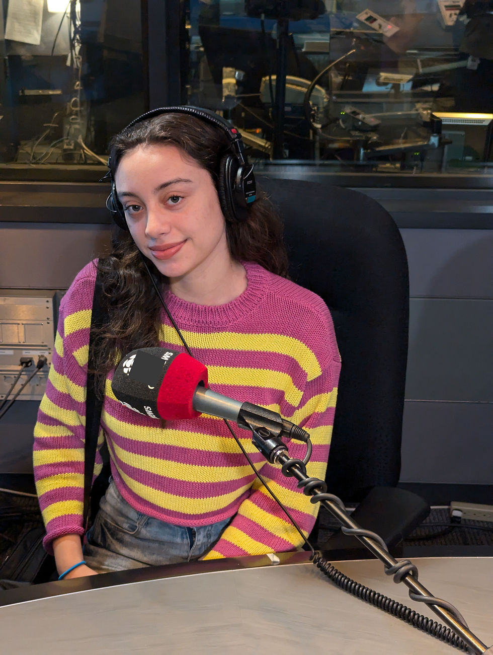 Young woman in a recording studio wearing headphones and a purple-yellow striped sweater, seated at a desk with a microphone. Calm expression.