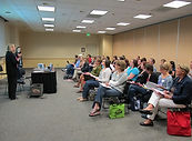A large group of people watches someone presenting in a conference room