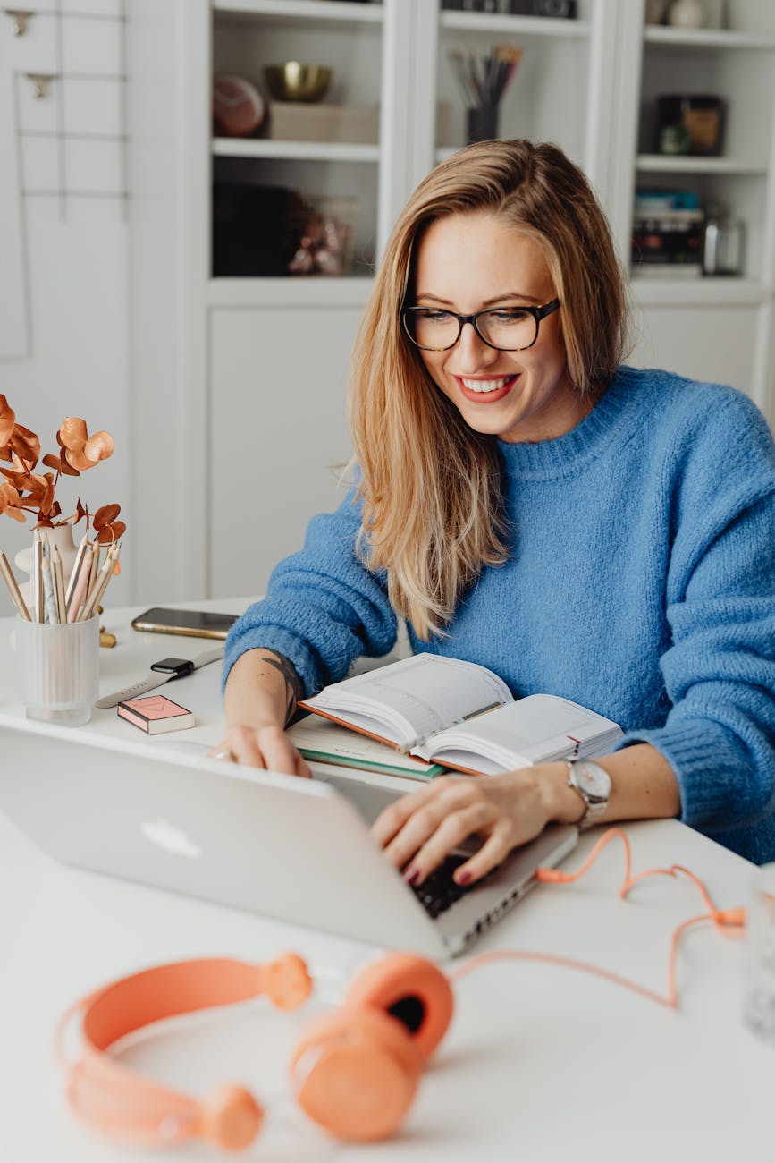 Uma mulher sorridente com óculos e cabelo loiro, usa um suéter azul, trabalha em um laptop em uma mesa organizada. Ao seu redor, há livros abertos, fones de ouvido cor de rosa, um smartphone, um relógio de pulso, um pote com lápis e flores secas. No fundo, uma estante branca com itens decorativos. Palavras-chave: mulher sorridente, óculos, suéter azul, laptop, mesa de trabalho, livros, fones de ouvido, smartphone, relógio, flores secas, estante branca, decoração.