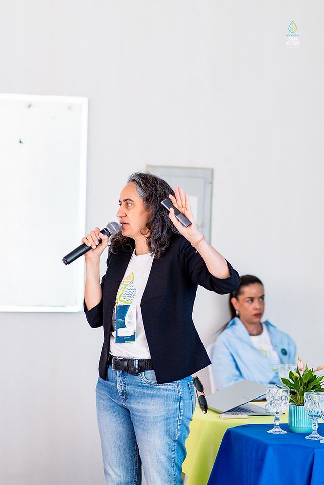 Fabiane Sebaio palestrante falando ao microfone em apresentação do evento, com projetor e mesa de apoio ao f