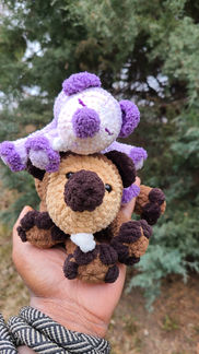 small crochet purple sleepy beaver on top of a brown sleepy baby beaver in a hand in front of pine trees in the background