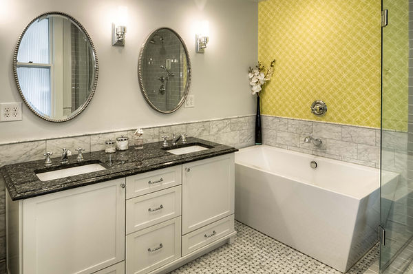A primary bathroom features a vanity with two sinks and dark grey countertops. Above the sinks are two circular, silver-lined mirrors. Next to the vanity is a rectangular, white, modern bathtub. The wall behind the bathtub faucet has green, geometric wallpaper.