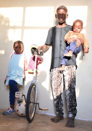 A brother and sister stand against a wall with a unicycle