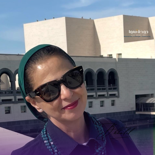 Woman in sunglasses smiles outdoors, wearing a green headband and blue beads. Beige building with arched windows in the background.