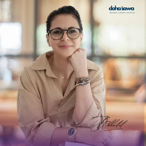 Smiling woman with glasses, beige shirt, and bracelets, sitting indoors. Warm blurred background. Text: "doha sawan" and signature "Behold."