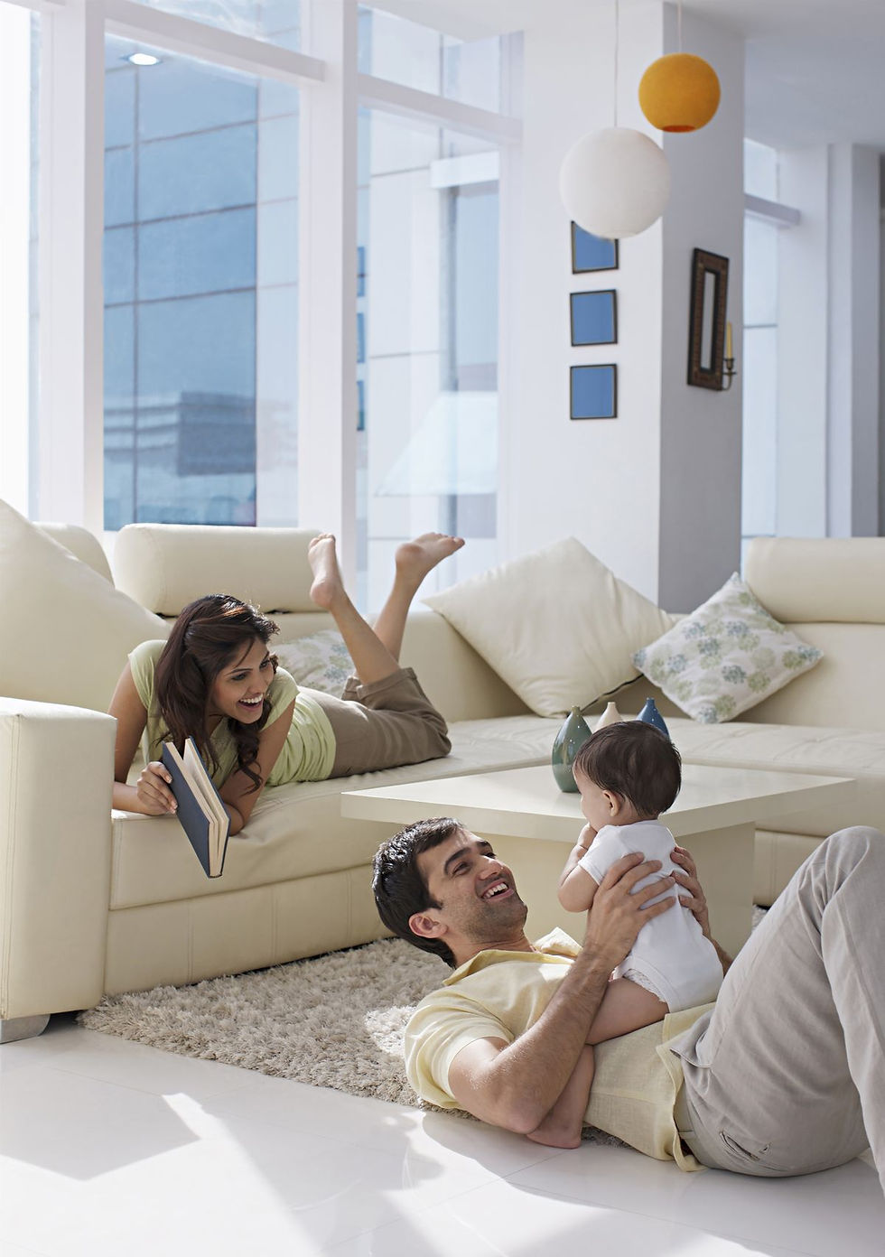 Couple smiling while moving boxes in a modern living room with large windows and a white couch. Bright atmosphere, unpacked boxes nearby.