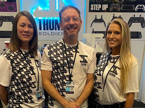 Tre membri del team Thumb Soldiers posano sorridendo allo stand fieristico del brand, circondati da pannelli espositivi con accessori e grafiche del marchio.