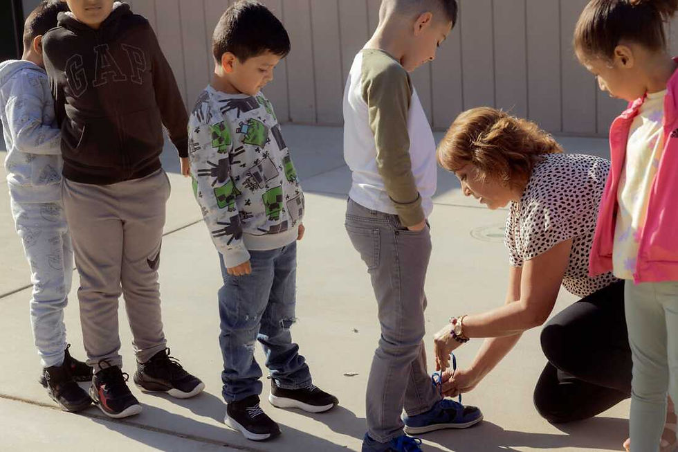 A preschool teacher kneels to help a child tie their shoes while other children wait in line, illustrating Finland’s gentle approach to teaching independence through patience and guidance in everyday routines.”