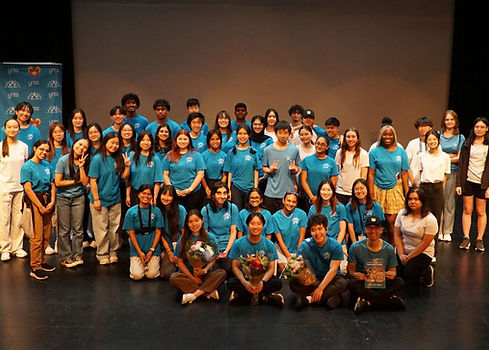 A large group of diverse young people in blue shirts posing on stage for a group photo, some holding flowers and a program booklet, likely after a performance or event.