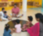 a group of children between the age of 4-10 sitting in the hall of a classroom with their teachers and talking.