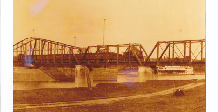 Train crossing truss bridge with steam boat in background at Red River in Fulton.