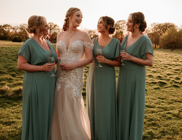The bride and her bridesmaids enjoy a candid moment at sunset in the scenic fields of Brookfield Barn. The bridal party is dressed in sage green gowns, holding glasses of wine. A beautiful example of relaxed bridal party photography and storytelling wedding imagery.