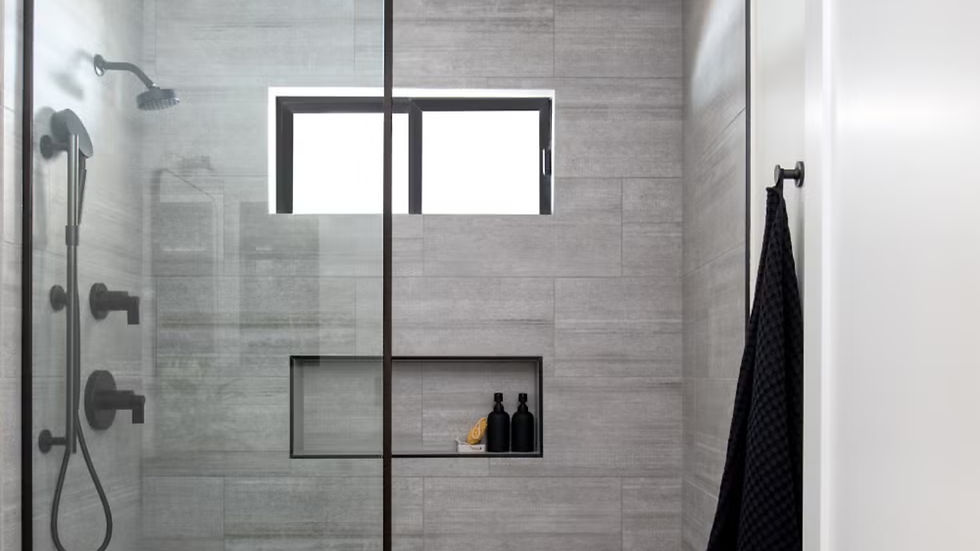 Modern gray tiled shower with glass door, black fixtures, inset shelf holding black bottles and soap. Towel on a hook; minimalistic vibe.