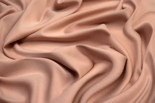 Smooth peach Rayon fabric, intricately folded, fills the frame. The texture is soft and flowing, creating a serene and elegant mood.