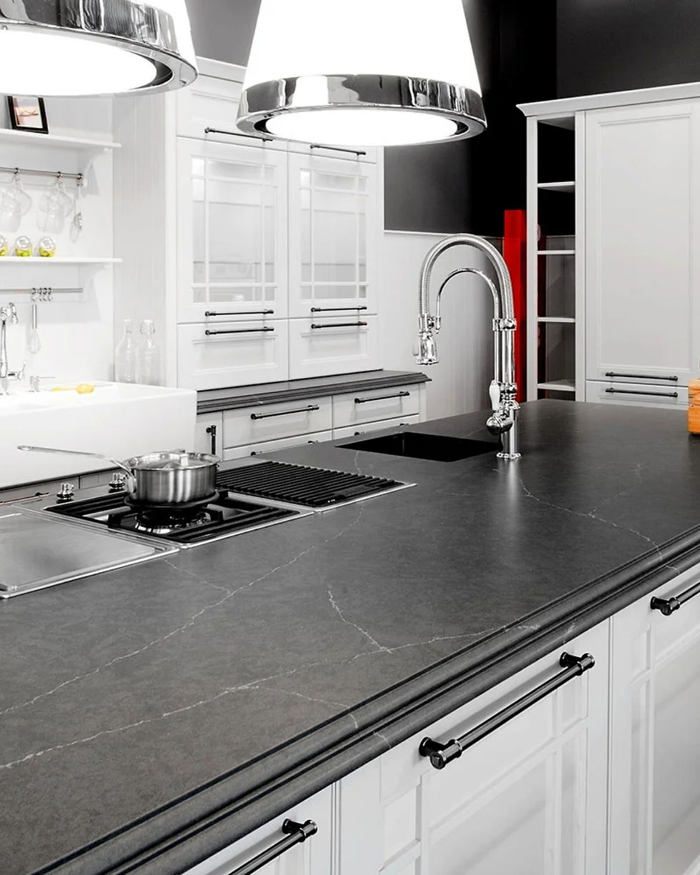 Modern kitchen with a sleek soapstone countertop and black faucet. Bright overhead lights, white cabinets, pot on stove, and subtle decor.