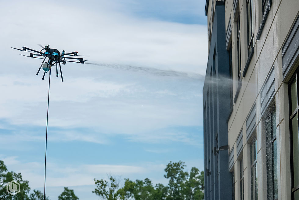 An industrial-grade exterior cleaning UAV, such as the Lucid Bots Sherpa, spraying a commercial building facade, demonstrating a safe, ground-based aerial positioning system for automated facility maintenance.