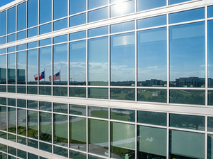 Tall, all glass corporate office building with clean, sparkling windows. In the reflection is a Texas and American flag waving in the breeze.